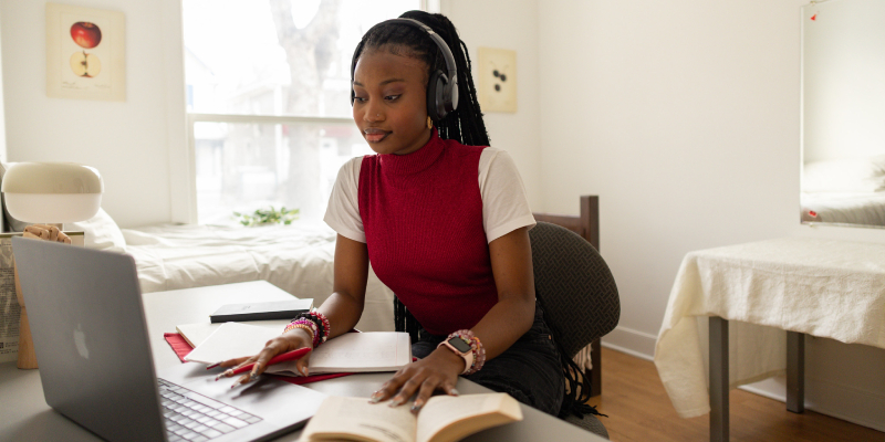 A student sitting at a laptop with an open book by one hand