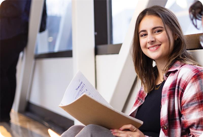 Student sitting in hallway holding a notebook and smiling at the camera