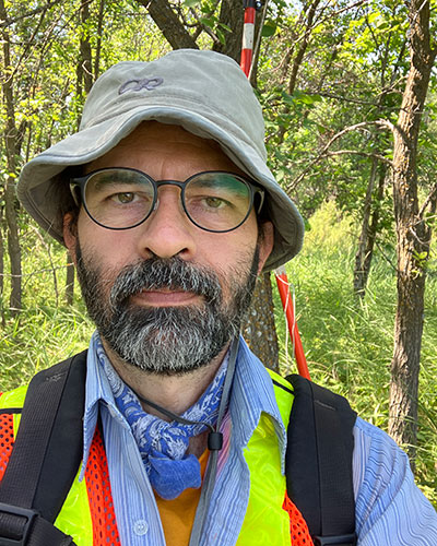 Germán Ávila-Sákar doing research in the field. The background looks like a forest and Germán is wearing a hand and a hankerchief around the neck.