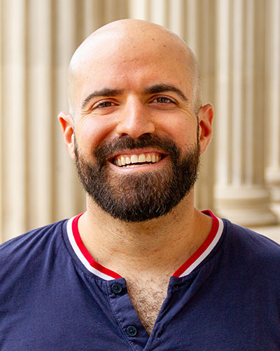 A headshot of Roman Belli. Roman is wearing a navy blue shirt with a red and white striped collar. There are pillars in the background.