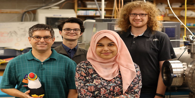 The photo is a group shot of Dr. Russell Mammei, Thomas Hepworth, Abeer Zahra, and Rylen De Vries standing in a research lab staring directly at the camera