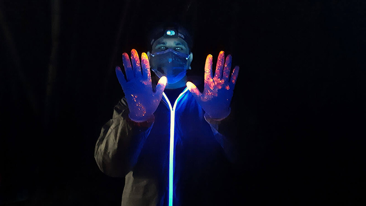 A person in a mask and plastic protective suit is covered in fluorescent powder, which is glowing under an ultraviolet light.