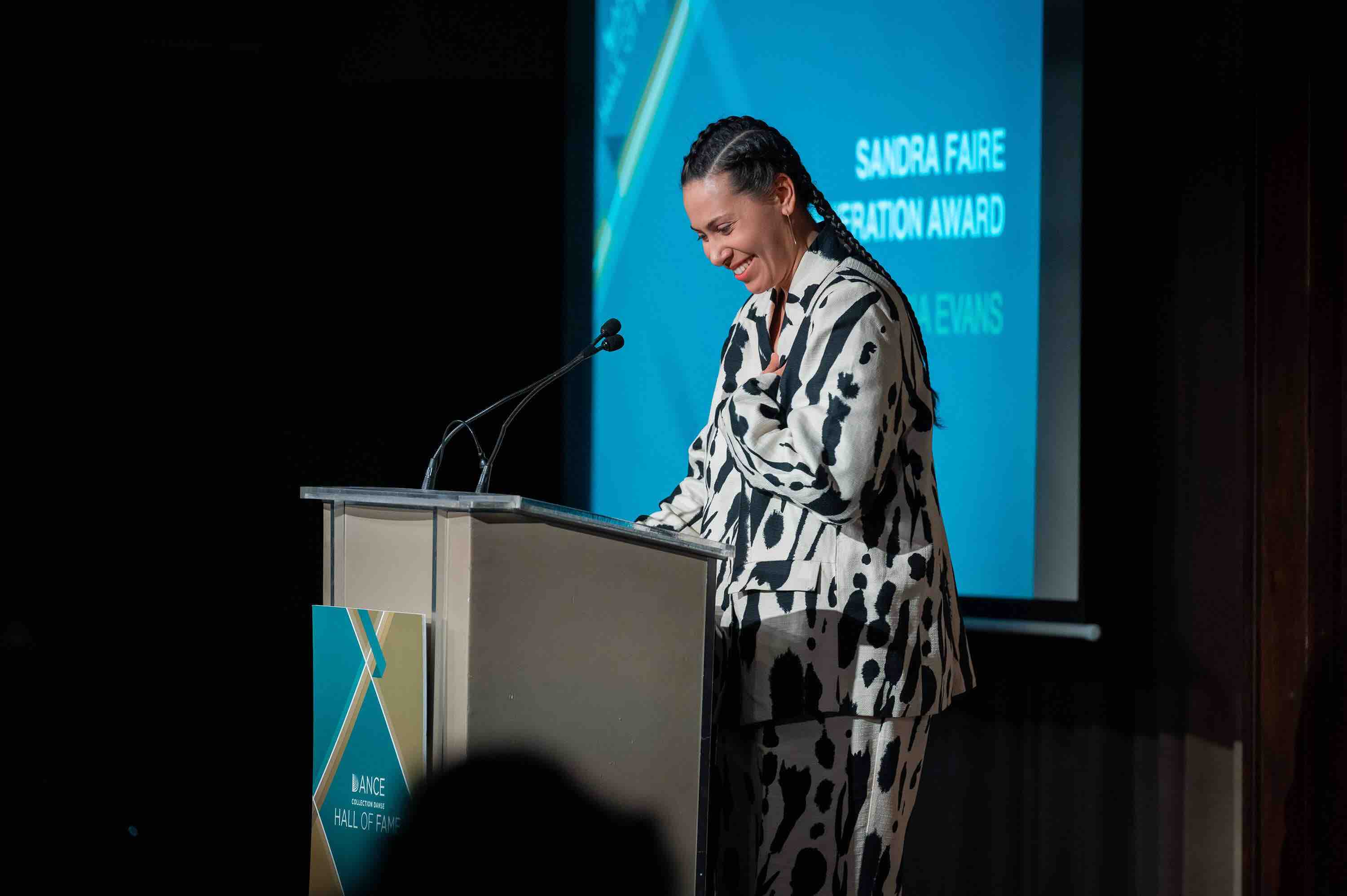 Aria Evans stands at the front of an audience accepting the DCD award. They are in a black and white paint blotch suit and their hair is braided. 