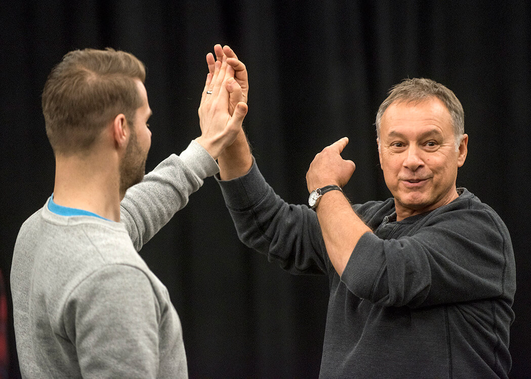 I’m standing in front of an actor, demonstrating and explaining something with an expressive gesture. My hand is raised, lightly meeting theirs in a kind of interactive movement, and I’m pointing with my other hand to emphasize my instructions. I’m wearing a dark sweater, and my focus is on engaging the person in front of me. Behind us, the background is blurred and dark, like a rehearsal or workshop space.